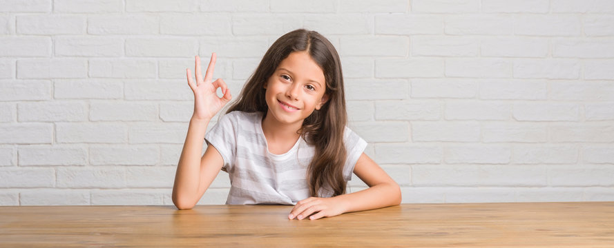 Young Hispanic Kid Sitting On The Table At Home Smiling Positive Doing Ok Sign With Hand And Fingers. Successful Expression.