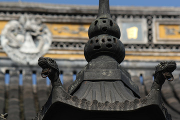 Top of an incence bowl with dragon images in front of a decorated roof top with a stone relief