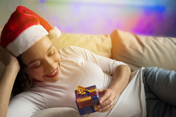 Happy African American woman wearing Santa hat looking at christmas gift lying in sofa at home. Blurred background with lights.