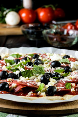 Raw uncooked pizza with pepperoni salami, black olives, basil and cheese.