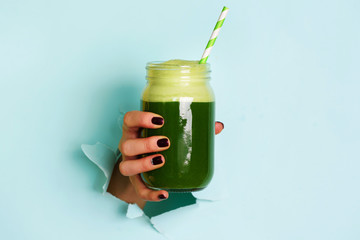 Woman hand holding glass jar of green smoothie, fresh juice against blue background. Healthy beverage, vegan, vegetarian concept. Banner with copy space