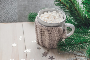 Obraz na płótnie Canvas White cup with marshmallows in a warm knitted jacket on a light wooden background with a Christmas tree and silvery stars.