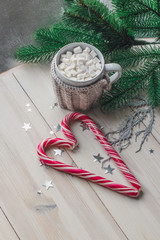 cup in a knitted jacket with two red caramel Christmas candies that lie in the shape of a heart on a white wooden background.