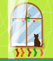 A cat sits on the windowsill by the winter and snow-covered window and looks out the window behind which it is snowing, the window with the curtain is dressed up with a garland and Christmas socks. Fl