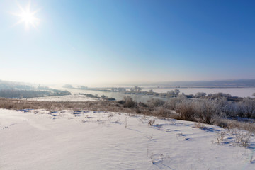 Fototapeta na wymiar Bright sun with a haze in the blue sky illuminates the river and snow-covered fields in the frosty morning