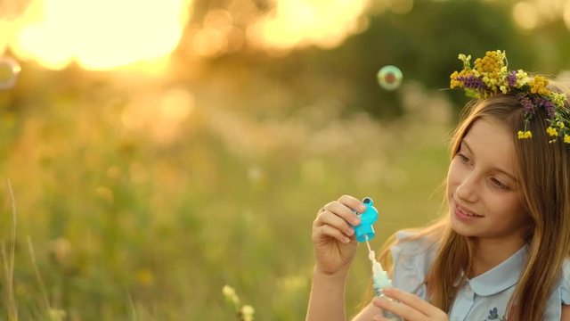 Girl in wreath is blowing bubbles on the meadow