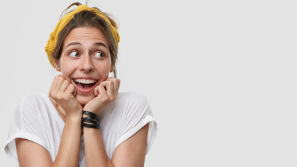 Glad positive European lady keeps both hands under chin, looks happily and mysteriously aside, dressed in casual outfit, has bracelet, stands over white background with copy space for your text