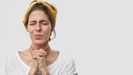 Photo of intense attractive European woman puts all efforts in pray, beggs for help, closes eyes,...