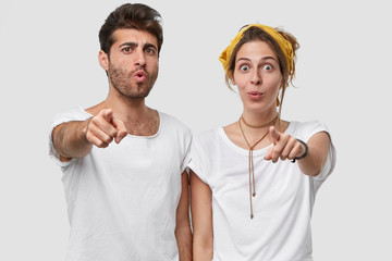 Horizotal view of emotive surprised Caucasian unshaven man and lady point directly at camera with index fingers, have astonished facial expression, notice something unbelievable, stand indoor