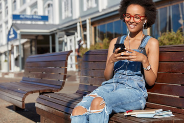 Photo of cheerful black woman holds cellular, types text messages, uses earphones, listens music, dressed in ragged overalls, models outdoor, enjoys playlist. Modern technologies, online communication