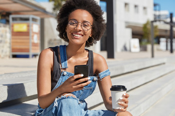 Young positive dark skinned teenager messages online, enjoys takeaway coffee from disposable cup, wears ragged denim overalls, enjoys spare time in open air. Street style. Lifestyle concept.