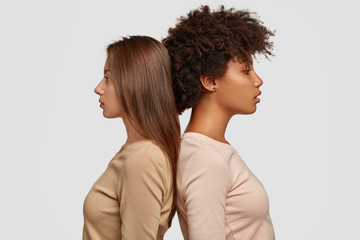 Two mixed race women stand in profile back to each other, have thoughtful expressions, dressed in casual clothes, think about cooperation, isolated over white background. Studio shot. Ethnicity