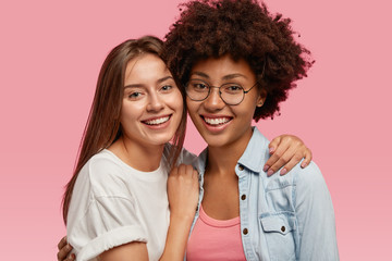 Portrait of friendly multiethnic women smile broadly, cuddle and enjoy togetherness, pose for family portrait, wear casual clothes, isolated over pink background. Ethnicity and friendship concept