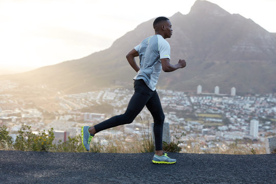 Self determined dark skinned sporty male runner wears sportclothes, runs on long distance across mountain road, enjoys fresh air, feels energetic and motivated. People, lifestyle and sport concept