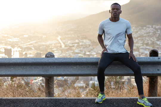 Healthy man with black skin, rests at road sign, wears casual clothes, looks confidently at camera, models against panoramic view with mountains and city, leads healthy lifestyle. Physical training