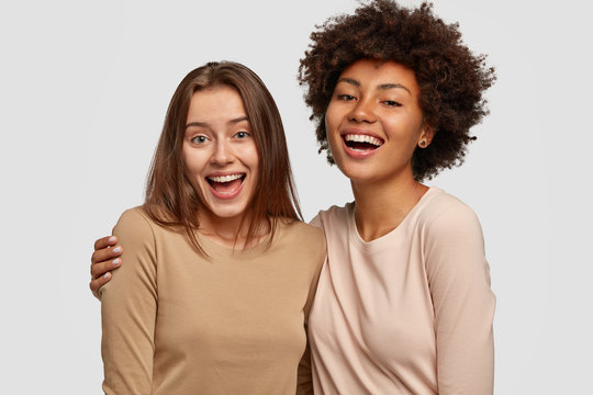Photo of joyful ladies embrace and enjoy togetherness, being of different of races, dressed in casual jumpers, isolated over white background. Diverse women laugh happily, have fun together.