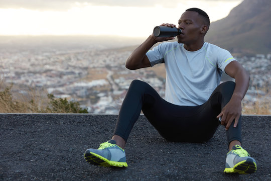 African American man drinks fresh water from bottle, rests on asphalt, sits against mountain background outdoor, feels relaxed, dressed in casual t shirt, sneakes and trousers. Relaxation concept