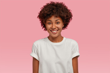 Indoor shot of attractive young African American woman with toothy tender smile, shows white teeth,...