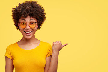 Joyful dark skinned woman with Afro haircut, points aside with thumb, pleased by big sale prices, wears sunglasses and casual t shirt, models over yellow background, shows copy space for your advert