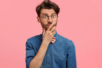 Photo of surprised frightened Caucasian bearded man keeps hand on mouth, looks with eyes full of shock, wears stylish denim shirt, notices something unbelievable, models against pink studio wall