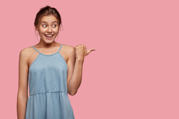Horizontal view of pretty woman with happy expression, dressed in blue dress, points with thumb on right side, shows free space against pink background, being in good mood, advertises item indoor