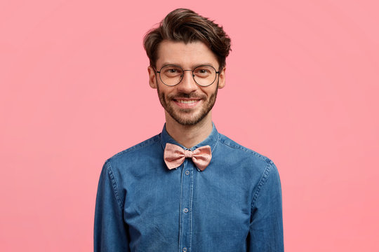 Photo of attractive smiling man with trendy hairstyle, positive look, dressed in fashionable festive outfit, stands against pink wall, rejoices good moments in life, prepares for date with girlfriend