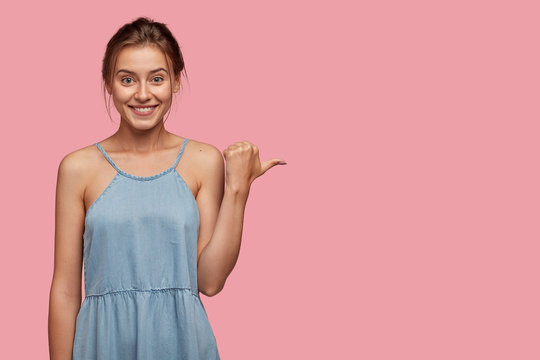 Studio shot of pretty Caucasian woman with glad facial expression, points with thumb on blank space, wears casual summer dress, shows place where she bought it, isolated over pink background