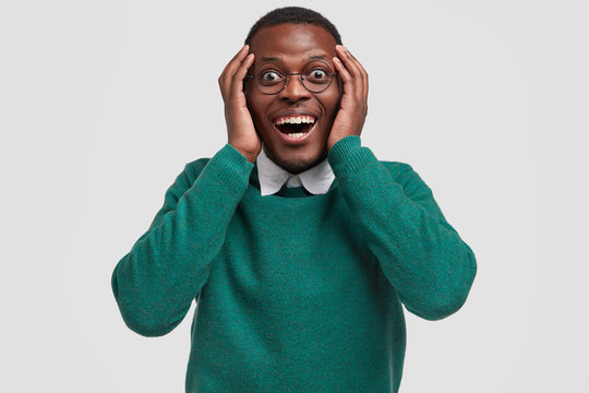 Positive young African American guy feels overjoyed as recieves money reward for good diligent job, keeps hands on head, smiles broadly, wears casual green jumper, isolated over white background