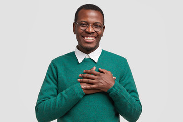 Handsome cheerful black man keeps both hands on chest, feels touched or thankful, smiles broadly,...