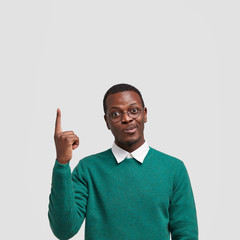 Photo of indecisive black male purses lips, lifts fore finger upwards, stands over white wall, wears sweater and shirt, shows free space for your advertisement or promotional content. Listen up