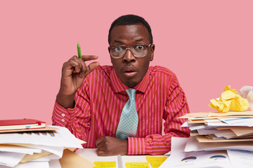 Photo of stunned displeased man makes size gesture, demonstrates how much attention and help he needs, completes task, models at desktop, has much work with financial project, wears tie and glasses