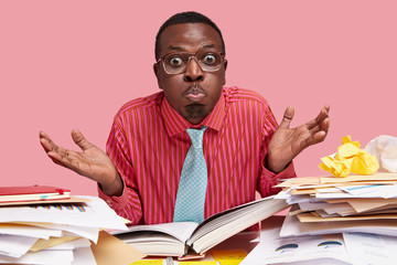 Funny puzzled black male wonk spreads palms with hesitation, blows cheeks and makes griamce, has fun as being tired of cramming material, has hard working day, sits at table with opened book, papers