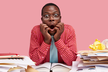 Bugged eyed young male with scared facial expression, opens mouth in bewilderment, keeps both hands on face, dressed in formal shirt, stupefied with much paperwork, isolated over pink studio wall