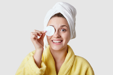 Joyful young woman removes make up with cotton disk, has toothy smile, uses cosmetics, wears warm yellow robe, isolated over white background. Pretty lady uses cotton pads, cares of her skin.