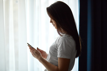 Sideways shot of dark haired young woman in white t shirt, checks email, enjoys free time at home, stands near window, uses modern cell phone, connected to wireless internet, sends text message