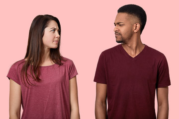 Obraz premium Sullen displeased multiethnic lady and male looks with discontent at each other, frown faces, dont like something, wear t shirt, model over pink background. People, feeling and relationship concept