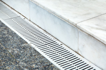 Obraz premium The gutter of the drainage system on the sidewalk, close-up metal grille light color on the background of marble tiles.