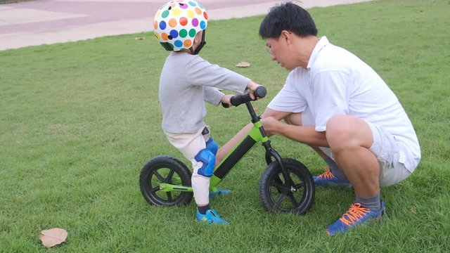 Father put protective knee pads on  Asian 2 year old toddler baby boy child, Dad and son having fun with balance bike (run bike) on at the park, Dad tech son to ride bike, father's day concept