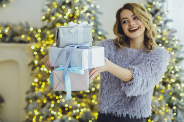 Young beautiful blonde woman in a gray sweater with a gift on hands near the New Year, Christmas tree with gifts