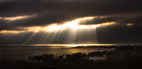 Beautiful dramatic yellow sun rays shine through a crack in cumulus clouds over Lake Michigan water and shoreline near Foster beach in winter in Chicago making a fantastic nature panorama background.