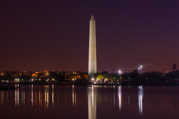 City skyline and Washington Monument at night during cherry blossom festival in Washington DC, USA. Colorful reflections of the monument and urban buildings in the water.