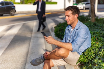 Mobile phone happy young man texting sms text message on smartphone waiting for meeting on city street. Urban lifestyle people outdoor in summer.