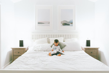 A little boy reading in his parents bedroom in his pyjamas.