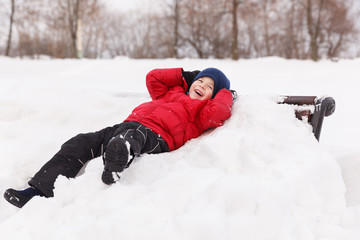 Fototapeta na wymiar Cute kid boy in a red jacket lying on the snow and laughing merrily. Happy child in a winter park outdoors. The concept of holiday and lifestyle
