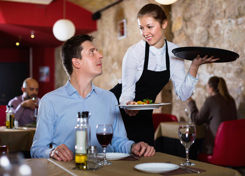 Polite waitress serving ordered dishes to smiling man at restaurant