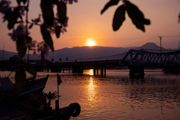 Sunset landscape with still river and leaves. Romantic sunset in tropical country. Urban bridge and bay with old ship.