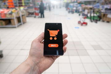 Shopping basket on a mobile phone screen. Button Buy. Hand holding mobile phone on Supermarket blur background.