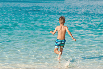 Active European boy in a striped swimming shorts is running towards the sea. He is having fun spending his holidays on the seashore.