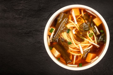 A bowl of miso soup with tofu, scallions, noodles, wakame, and enokitake mushrooms, shot from the top on a black background with a place for text