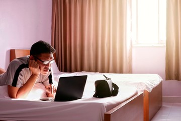 Mature Asian man using laptop computer to work in bed with flare light on window in hotel bedroom at morning, lifestyle and technology concept